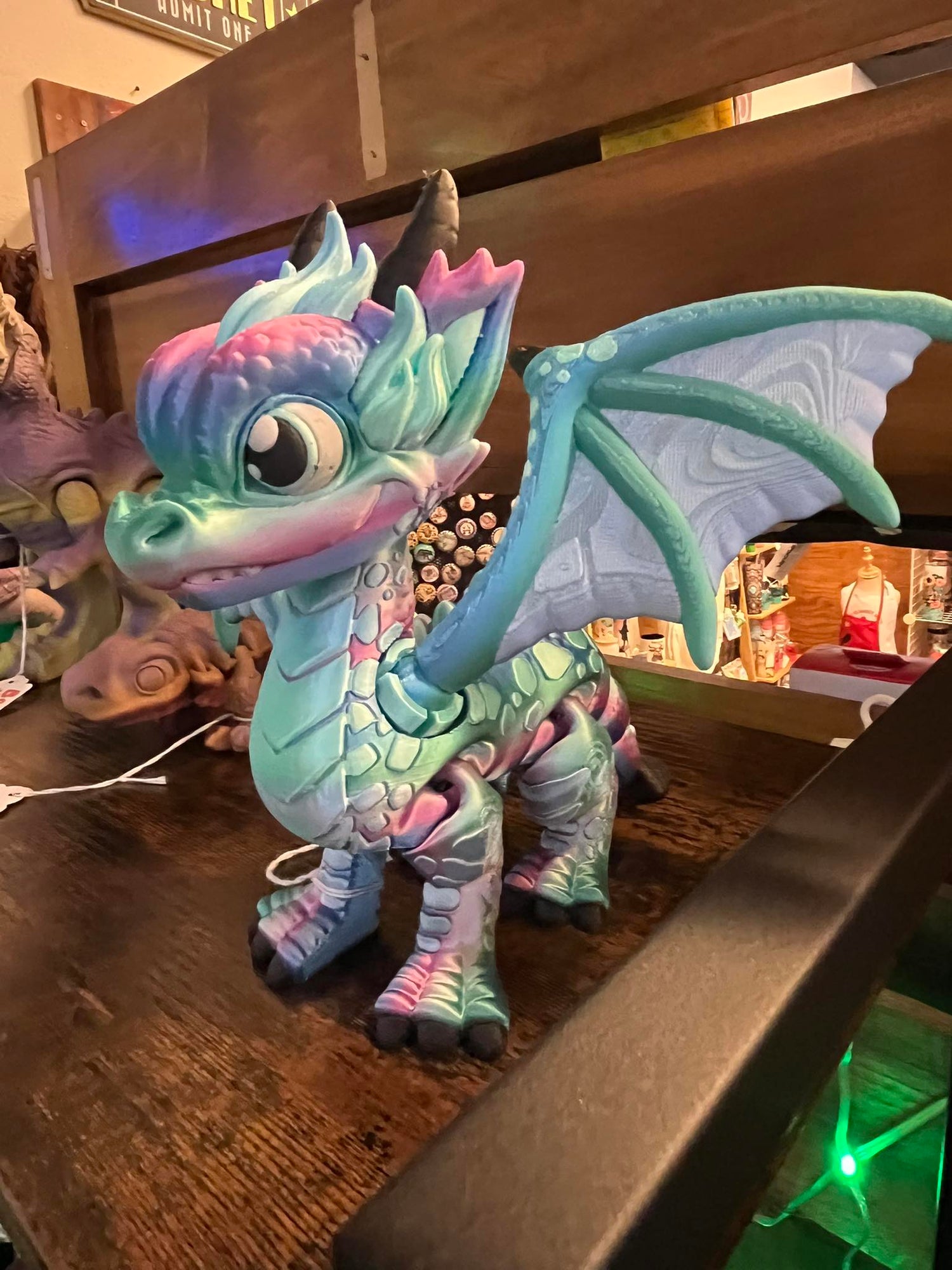 Colorful 3D printed dragon figurine with wings spread, featuring pastel shades of blue, pink, and green, displayed on a wooden shelf inside a shop.