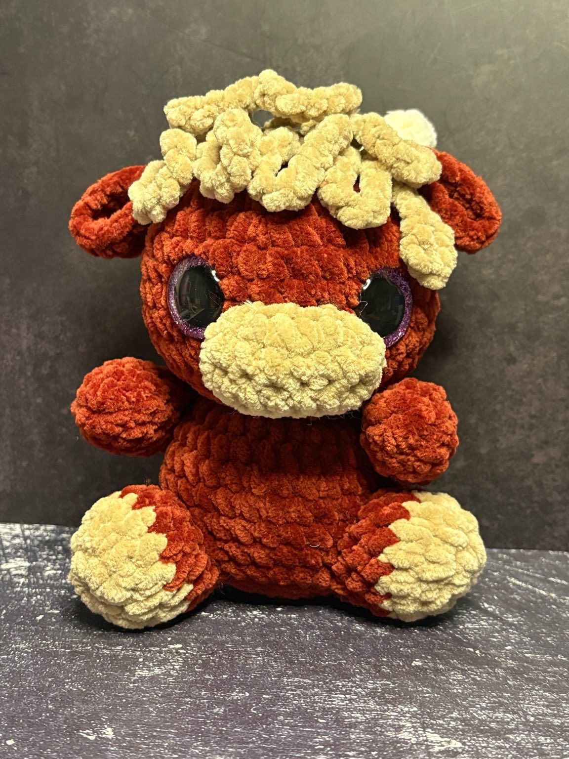 Hand-crocheted plush cow made with soft textured yarn in warm brown and cream colors, featuring stitched details and large shiny eyes, sitting against a neutral background.
