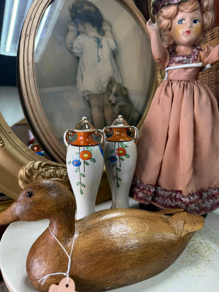 Vintage display featuring a carved wooden duck decoy, a pair of hand-painted floral vases, a framed vintage child portrait, and a dressed doll, arranged together on a shelf.