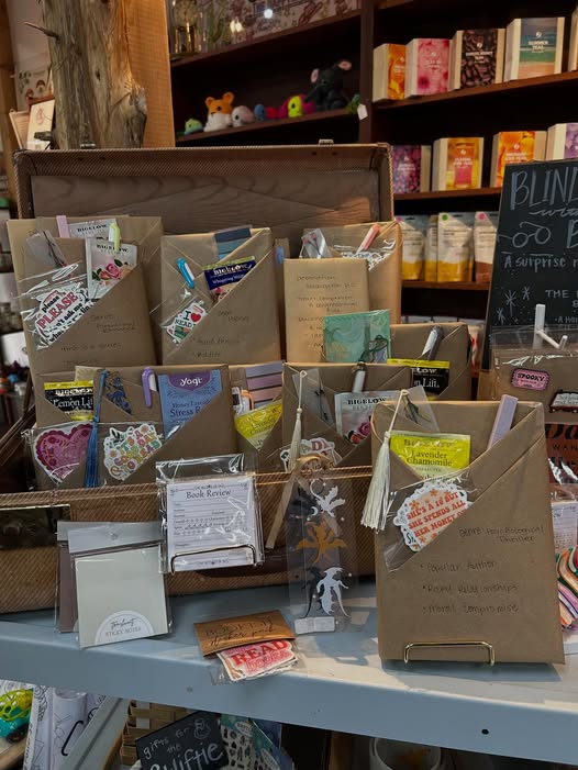 Wrapped “blind date with a book” packages displayed on a tabletop, each labeled with handwritten genre clues and paired with small reading-themed extras, arranged in front of shelves filled with books and gifts.