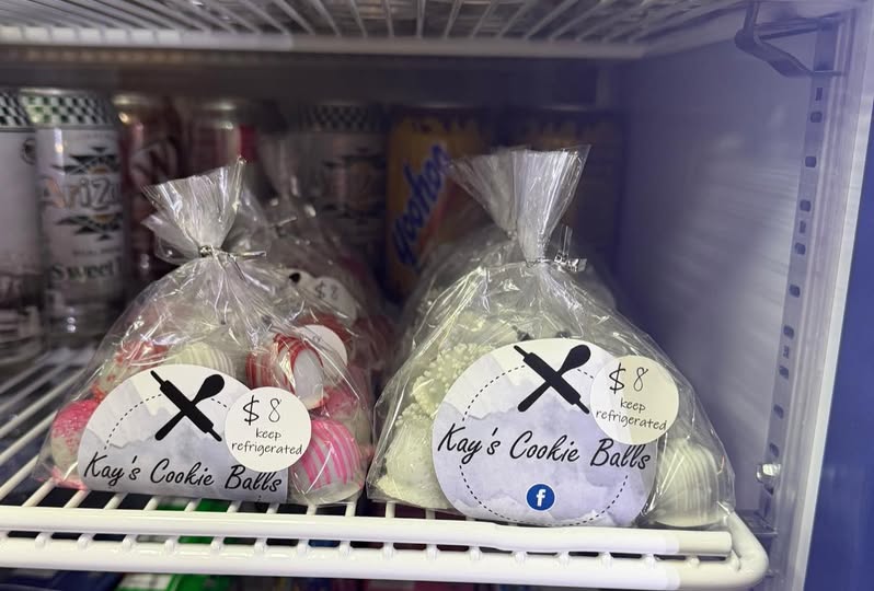 Handmade forged knives displayed on a wooden shelf, each set in a leather sheath and hung from a natural wood branch, with turquoise bead accents and small forged items arranged below.