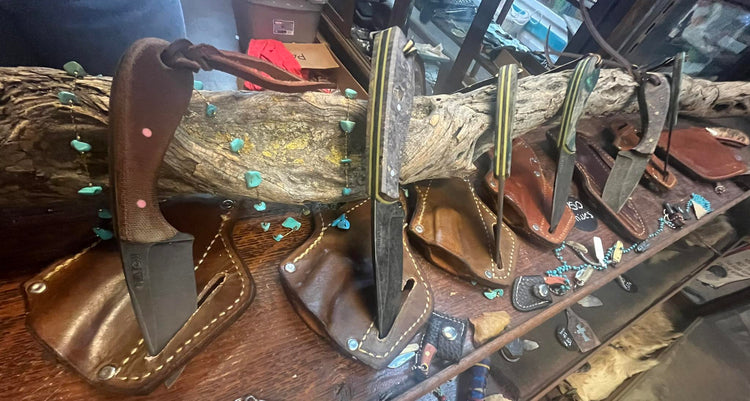 Handmade forged knives displayed on a wooden shelf, each set in a leather sheath and hung from a natural wood branch, with turquoise bead accents and small forged items arranged below.